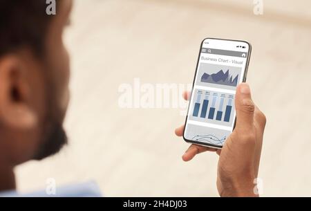 Millennial black guy with beard uses website or app with financial profit charts on smartphone screen Stock Photo