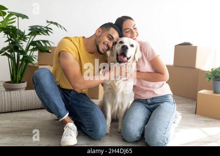 Happy young diverse couple hugging their cute dog, sitting on floor among carton boxes, moving to new home with pet Stock Photo