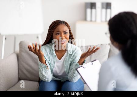 Stressed black woman sitting on couch, having consultation with psychologist, discussing her problem at modern clinic Stock Photo