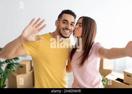 Lovely Caucasian woman kissing her Arab boyfriend, taking selfie, moving to new home together Stock Photo