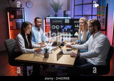 Multiracial team of four financiers having meeting at office room for discussing situation on trade market. Men and women using modern laptop and tablet for work. Cooperation concept. Stock Photo