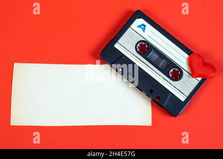 Old Audio Cassette with a Heart Shape and Blank Paper on the Red Background closeup Stock Photo