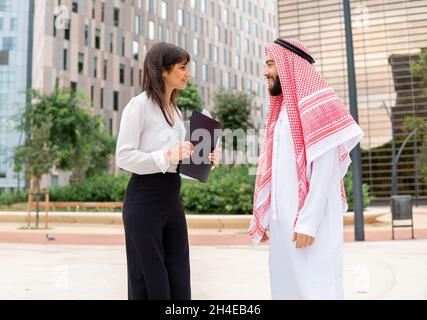 Friendly female manager with folder smiling and looking at bearded Arab male in traditional clothes during conversation on city street Stock Photo