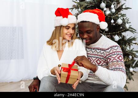 Joyful cozy moments in winter holidays. A multinational couple opens a Christmas present. A loving young black man and a white woman are sitting next Stock Photo