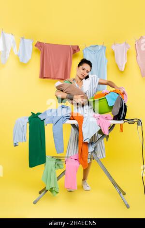 woman talking on smartphone while holding baby doll and laundry bowl near pile of clothes on ironing board on yellow Stock Photo