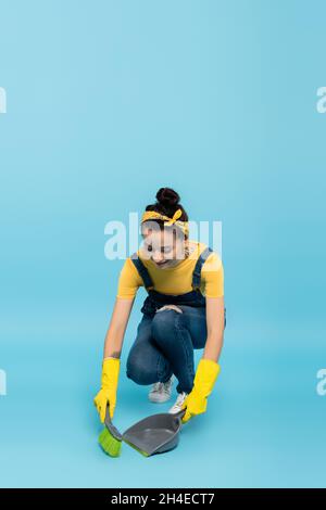 positive housewife in denim overalls and rubber gloves sweeping floor on blue Stock Photo