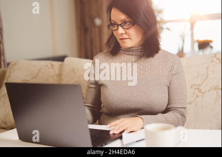 Working At Home. working remotely from home on laptop, sitting on couch in living room. businesswoman working on laptop Stock Photo