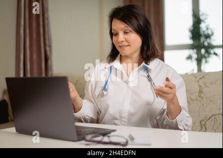 Female general practitioner wears white coat speaking videoconferencing on laptop computer using online video call consultation app. Remote medical he Stock Photo
