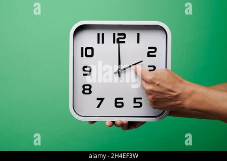 a young caucasian man sets a white clock backward, at the end of the summer time, on a green background Stock Photo