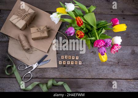 Happy Easter inscription in wooden letters. Gift wrapping. Mix of spring tulips flowers. Background with flowers tulips close-up different colors. Gift. Top view. Copy space. Craft papper. Stock Photo