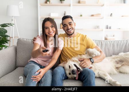 Happy young interracial couple sitting on couch with their funny dog and watching TV at home Stock Photo