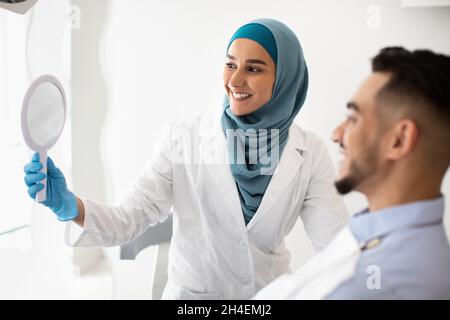 Muslim Dentist Woman Holding Mirror, Showing Teeth Treatment Result To Male Patient Stock Photo