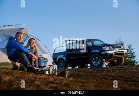 Young woman and man hikers sitting in tourist tent on grassy hill near black automobile. Loving couple travelers resting in camp tent under beautiful blue sky while hiking together in mountains. Stock Photo