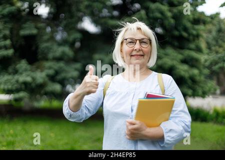 Smiling blonde teacher woman lady 40s 50s years old wearing eyeglasses standing hold notepads showing thumbs up looking camera outdoors Stock Photo