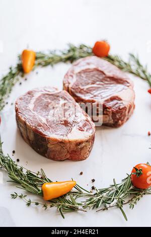 raw two ribeye steaks in the center of a Christmas wreath of rosemary thyme cherry tomatoes and bell peppers on a white marble surface. christmas dinn Stock Photo