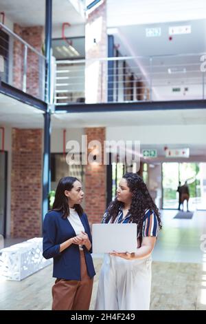 Two diverse businesswomen talking and using laptop in modern office with colleagues in background Stock Photo