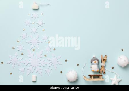 Winter Christmas composition. A toy penguin with skis rides a sleigh. The shape of a Christmas tree from snowflakes on a blue background. Top view fla Stock Photo