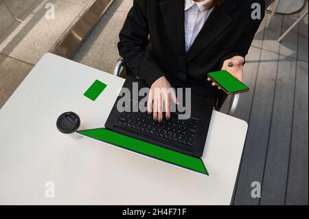 High angle view. Unrecognizable woman with smartphone with chroma key green screen, typing on laptop keyboard sitting at a table with coffee cup and g Stock Photo