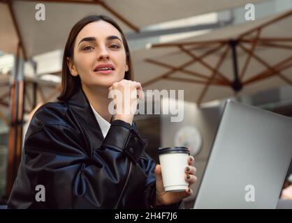 Lifestyle portrait of beautiful brunette woman, copywriter, freelancer with paper cup of takeaway coffee, smiling, looking at camera, working on lapto Stock Photo