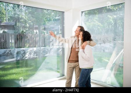 cheerful man pointing with hand at panoramic windows while hugging wife in new house Stock Photo