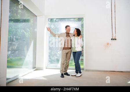 happy man pointing with hand at panoramic windows while hugging wife in new house Stock Photo