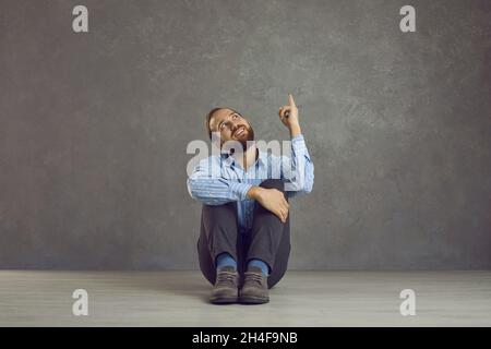 Funny caucasian man thinking sitting with raised finger on the floor on gray background. Stock Photo