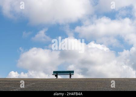 Low angle view of an empty park bench on top of one of the asphalt covered dikes in the Netherlands against a white clouded blue sky Stock Photo
