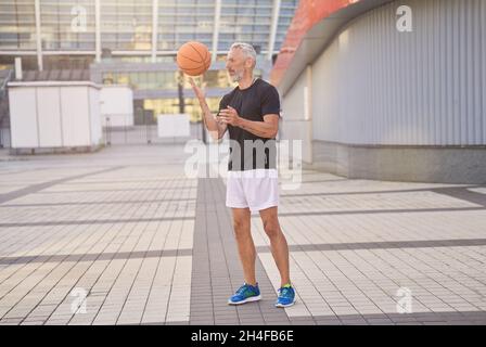 Full length shot of sportive mature man in sportswear standing outdoors and playing with basketball ball Stock Photo