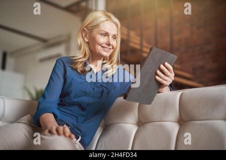 Relaxed mature woman smiling while reading e book using digital tablet pc, relaxing on a couch at home in modern apartment Stock Photo