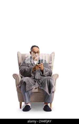 Mature man in a bathrobe sitting in an armchair and using a smartphone isolated on white background Stock Photo