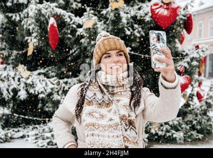 Portrait of a beautiful happy young woman taking a selfie in front of christmas tree on snowy day in city Stock Photo