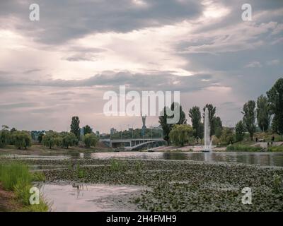 View over the Motherland monument, bridge and fountains in water canals of Rusanivsky district in Kyiv. Murky weather with cloudy sky at background. Stock Photo