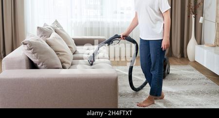 young woman in white shirt and jeans cleaning carpet under sofa with vacuum cleaner in living room, copy space. Housework, cleanig and chores concept Stock Photo