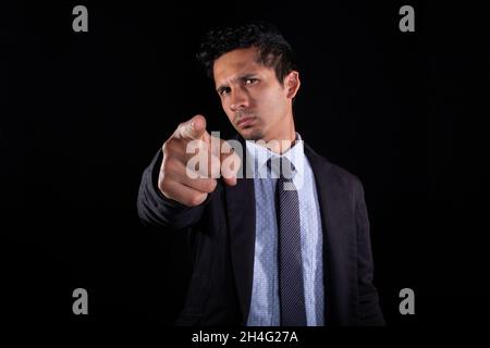 Businessman pointing forward with his finger. Young adult with serious and distrustful expression judging with finger. Isolated on black background. Stock Photo