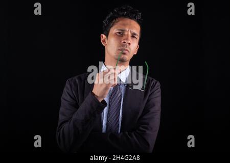 Very thoughtful businessman with glasses in his hand, isolated on black background. Concentrated young businessman looking straight ahead while Stock Photo