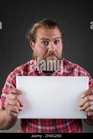 Man, worker in plaid shirt with beard and long hair with funny face expression holding small horizontal blank white board, in studio Stock Photo