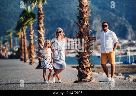 Happy family of mother father and young daughter stands under palm tree with rised hands. Beach holidays concept Stock Photo