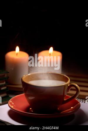 Hot yellow mug with drink, steam and two burning candles together on the table, black background space for text. Stock Photo