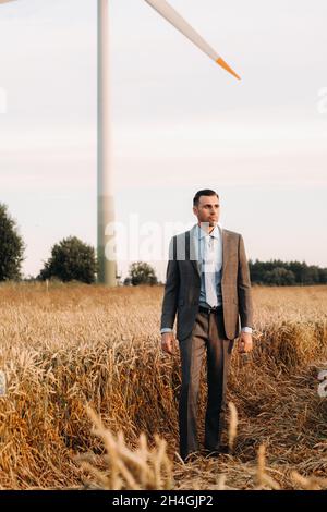 Portrait of a businessman in a gray suit on a wheat field against the background of a windmill and the evening sky. Stock Photo