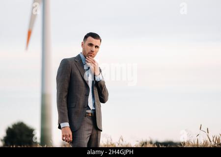 Portrait of a businessman in a gray suit on a wheat field against the background of a windmill and the evening sky. Stock Photo