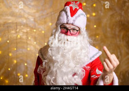 Portrait of an angry santa claus wearing glasses with white long beard showing middle finger marks against a wall background with garland. A man in costume of a fairytale hero shows a finger gesture Stock Photo