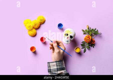 Woman with brush, paints and human skull on lilac background. Mexico's Day of the Dead (El Dia de Muertos) Stock Photo