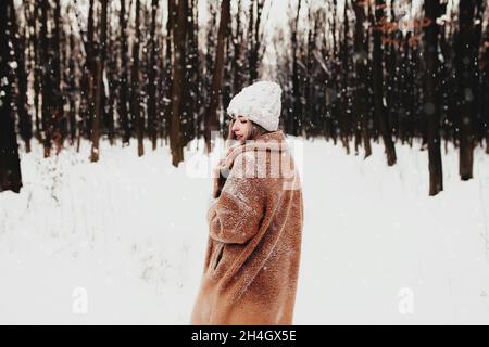 Photo of young beautiful woman in fur coat standing on the white snow in winter forest. Girl is smiling and happy with closed eyes. Female has winter Stock Photo