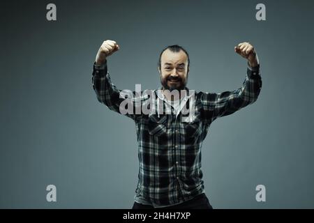 Half body portrait of bearded man in check shirt raising hands in triumph, studio background. Stock Photo