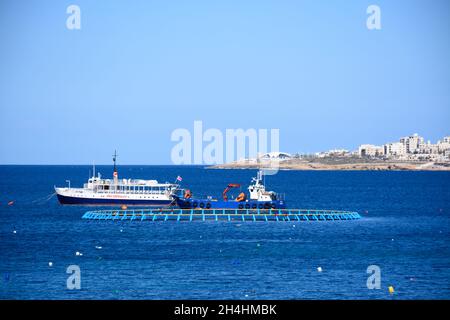 Fish farming off the coast with boats moored in the bay and views towards the town, Xenxija Bay, San Pawl, Malta, Europe. Stock Photo