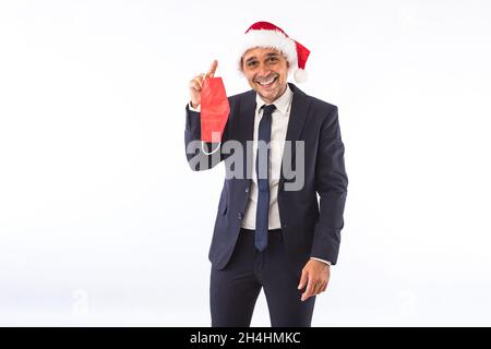 Businessman dressed in suit, tie and Christmas hat of Santa Claus, smiling and holding a red mask, on white background. Christmas celebration concept. Stock Photo