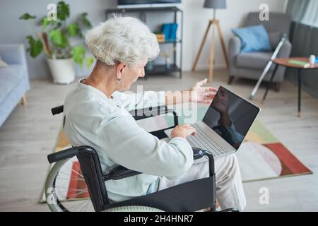 High angle portrait of white haired senior woman in wheelchair using laptop at home Stock Photo