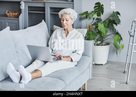 Full length portrait of modern senior woman using laptop while laying on couch at home, copy space Stock Photo