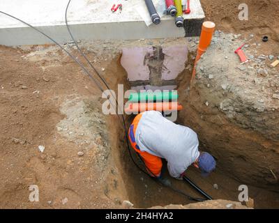 a fitter with orange pants is laying the telephone and data cables near other lines in the trench Stock Photo