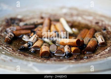 Viele Zigarettenstummel in einem grauen Standaschenbecher, draußen stehend, in den es hinein geregnet hat. Stock Photo